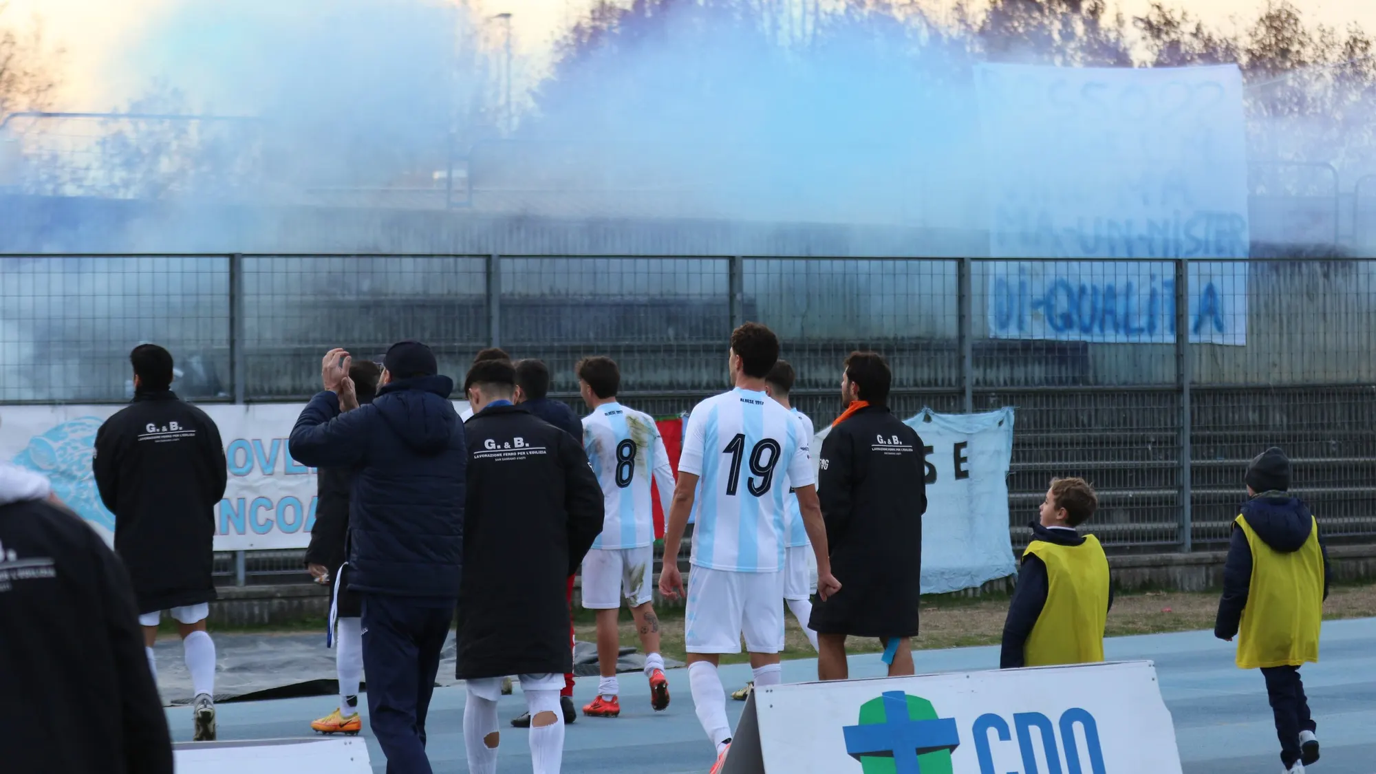 I giocatori dell'Albese rientrano verso la panchina mentre il fumo azzurro dei tifosi invade il campo