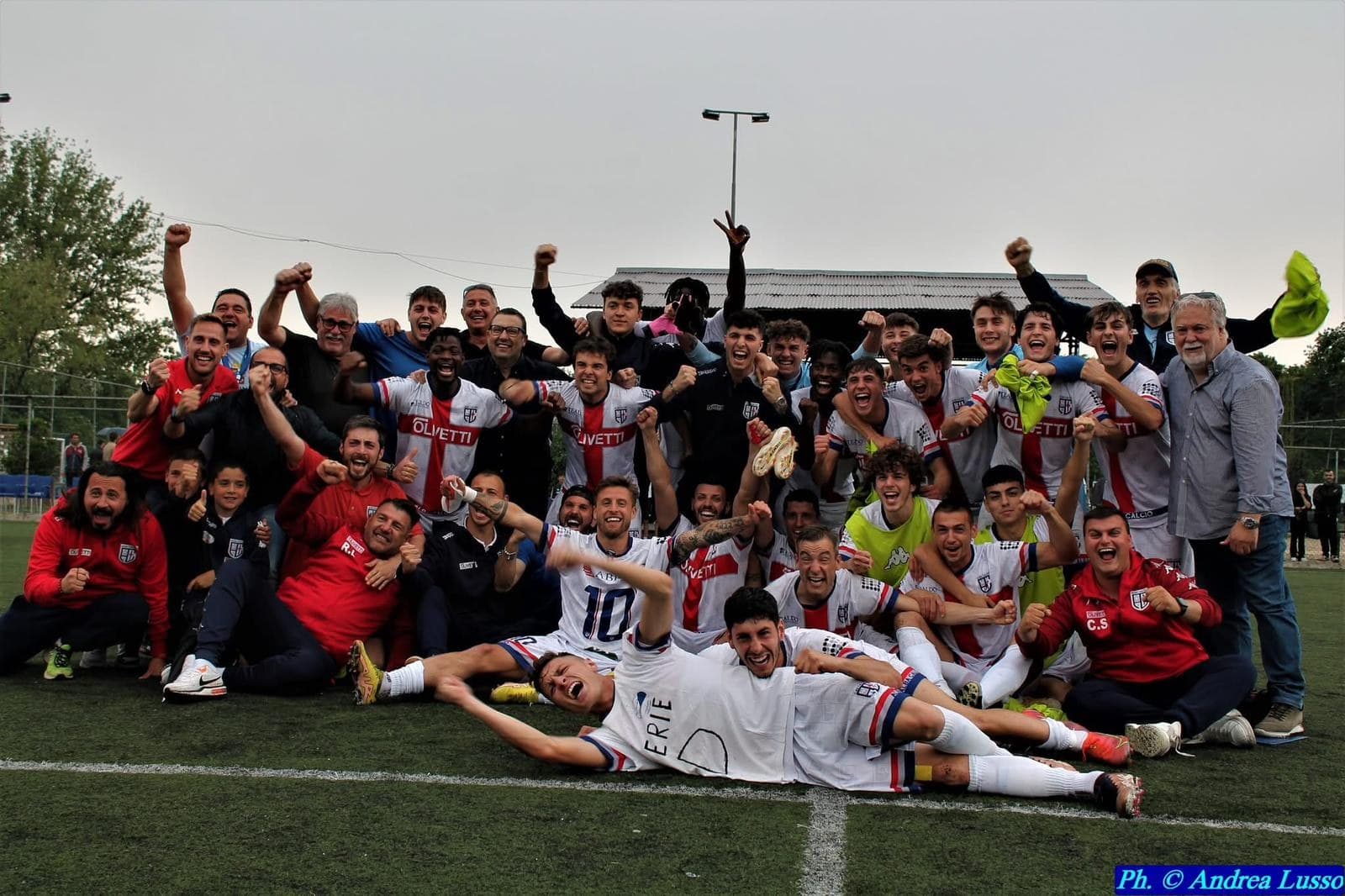 La squadra dell'Alba Calcio e il suo staff festeggiano in campo a braccia alzate, in maglia bianca con la croce rossa