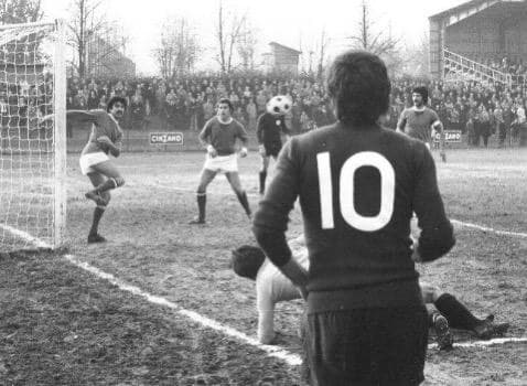 Azione di gioco in bianco e nero vista da dietro il numero 10: un attaccante calcia verso la porta davanti alla tribuna gremita