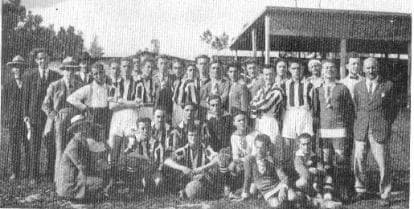 Fotografia d'epoca in bianco e nero: giocatori e dirigenti dell'Albese in posa a bordo campo davanti alla tribuna in legno